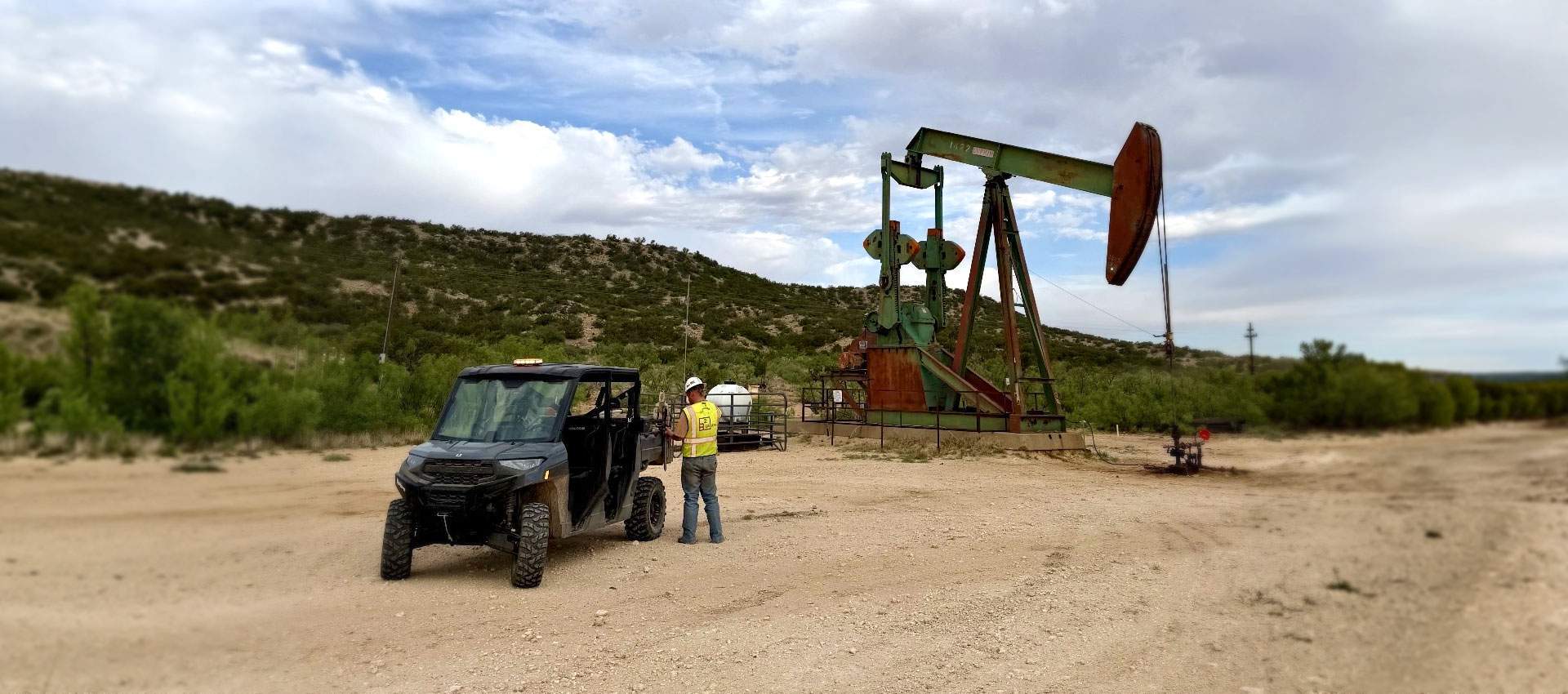 Crew member in a high-visibility vest at a pumpjack lease with a UTV parked on the caliche pad