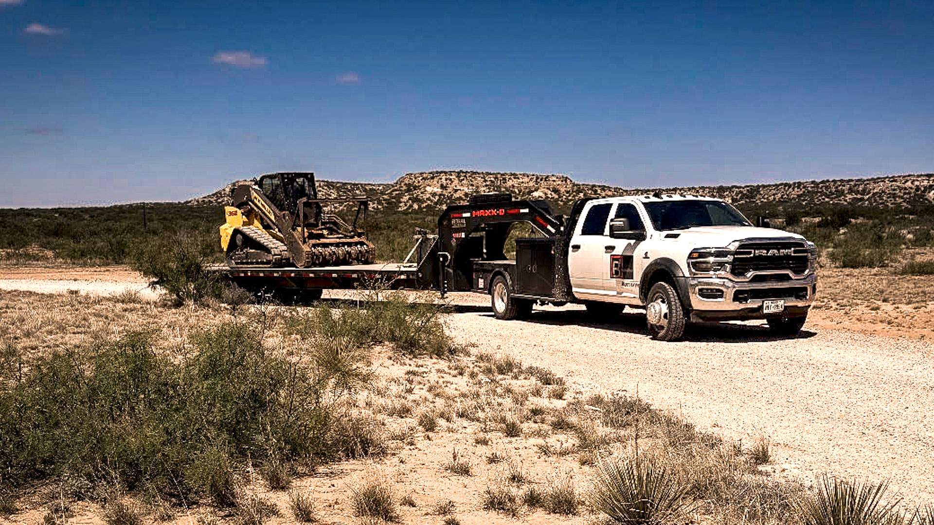 Flatbed truck hauling a tracked mulcher down a ranch two-track through desert scrub