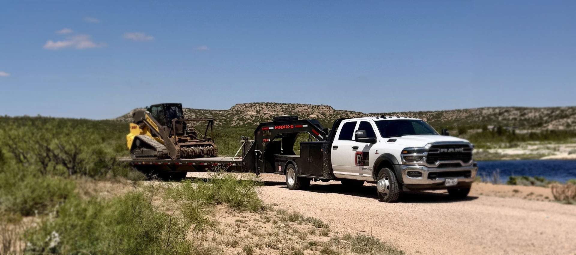 Crew-cab flatbed truck hauling a tracked mulcher on a gooseneck trailer along a rural roadside